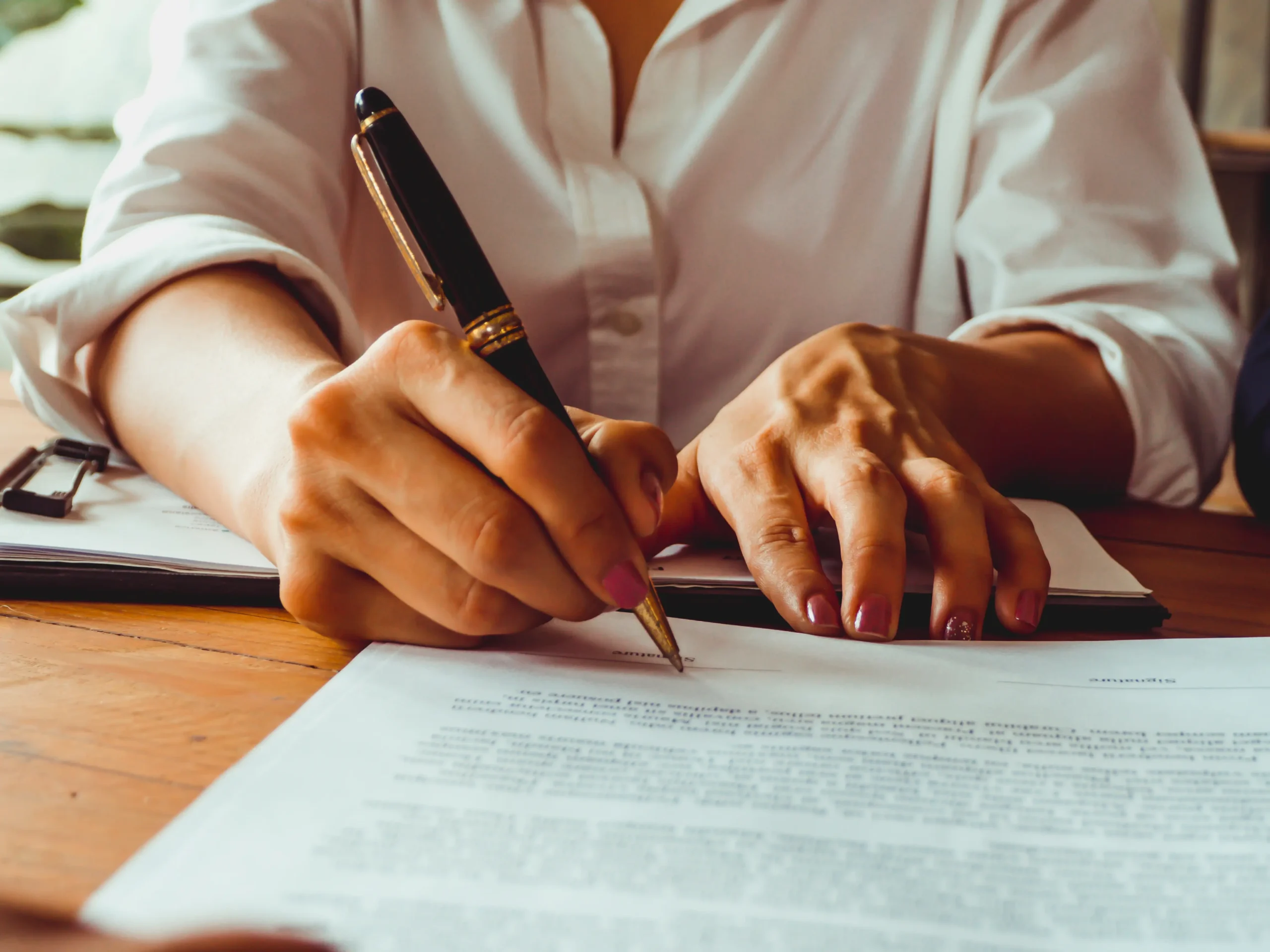 A letter sits on a table with a person signing it with a pen.